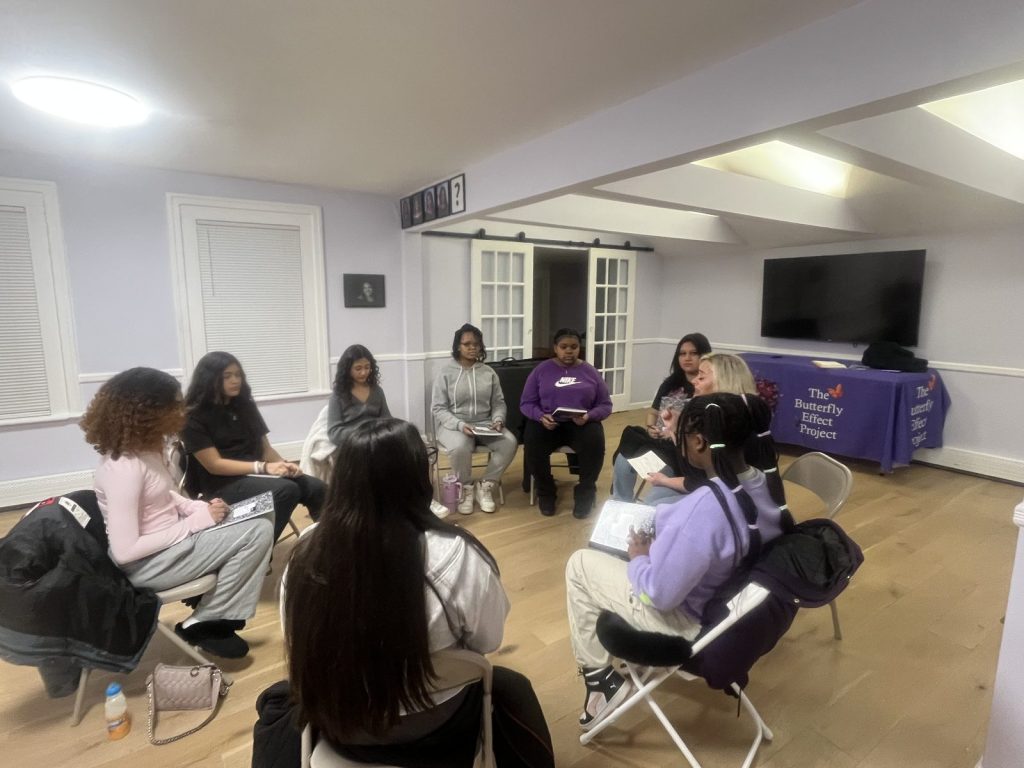 Young women sit in a circle of folding chairs in a bright room with Butterfly Effect table branding.